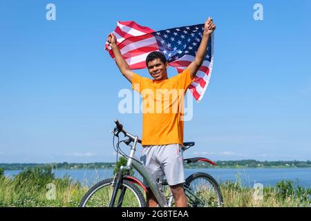 Afroamerikanischer Mann, der die USA-Flagge hält und winkt und mit dem Fahrrad steht Stockfoto