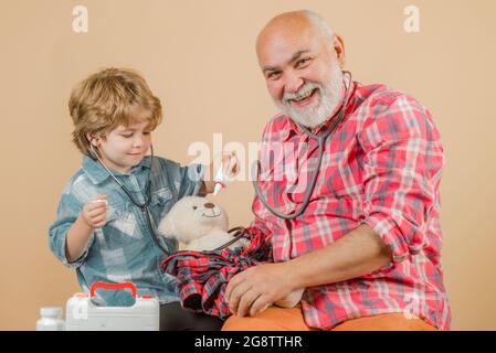 Kleiner Arzt. Charmanter kleiner Junge, der eine Wange seines Großvaters untersucht. Mann im bunten Strickschal mit Medizin. Älterer Mann mit kleinen niedlichen Stockfoto