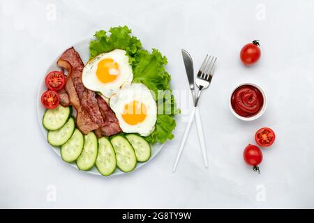 Gebratene Hühnereier mit Speck und Gemüse auf einem grauen Tisch. Frühstückskonzept. Ansicht von oben. Flache Lay-Komposition. Das Essen am Morgen. Fres Stockfoto