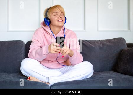 Eine junge Frau sitzt mit einem Tablet auf der Couch und hört Musik über Kopfhörer. Jugendkultur. Stockfoto