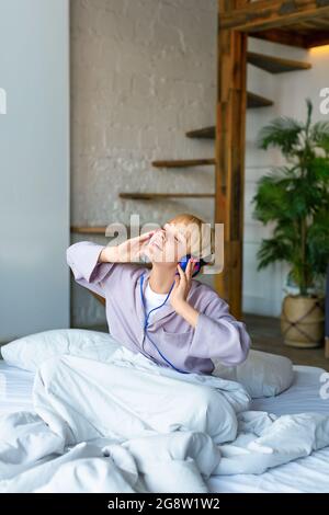 Eine Frau wachte auf und hörte Musik über Kopfhörer, während sie im Bett saß. Sie singt mit, während sie ihrem Lieblingslied zuhört. Moderne Technologie Stockfoto