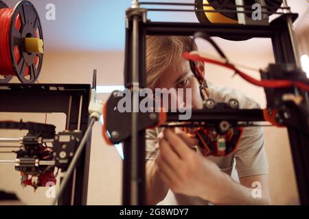 Junger männlicher Mechaniker, der den Mechanismus des modernen 3D-Druckers während der Arbeit in der Werkstatt repariert Stockfoto