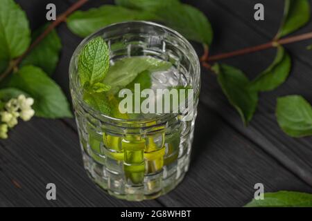 Erfrischender, kalter Mojito-Cocktail mit Limette und Minze im Glas auf verbranntem schwarzem Holzhintergrund, verziert mit Sommerblumen. Stockfoto