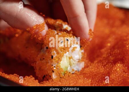 Nahaufnahme der Hände des Küchenchefs, die Sushi in fliegendem Fischrogen (Tobiko)-rotem Kaviar aufrollen - konzentrieren Sie sich auf die Rolle. Stockfoto