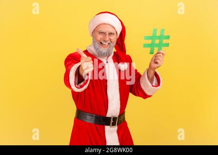 Lächelnder älterer Mann mit grauem Bart, trägt ein weihnachtsmann-Kostüm, hält einen grünen Hashtag in den Händen und zeigt den Daumen nach oben, empfohlene Website. innenstudi Stockfoto