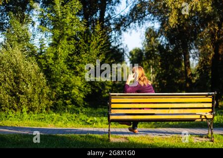 Unschärfe kaukasisch blonde Frau reden, sprechen am Telefon draußen, im Freien. 40-jährige Frau in lila Bluse im Park auf der Bank. Erwachsene Frauen Stockfoto