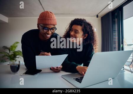 Nahaufnahme eines gemischten Rennpaares mit Laptop für Online-Banking. Glückliches afroamerikanisches Paar, das lächelt und einander anschaut, während es zusammenarbeitet Stockfoto
