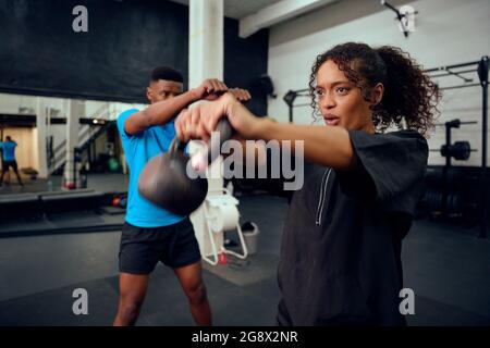 Afroamerikanischer männlicher Personal Trainer, der afroamerikanische Frauen mit einer Kettlebell-Routine im Fitnessstudio anweist. Gemischte Rennfreunde, die Cross fahren Stockfoto