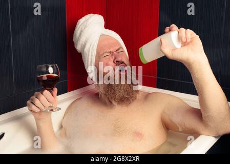Fröhlicher bärtiger Mann, der ein Schaumbad nimmt, ein Glas Rotwein hält und mit Gesichtsausdruck in eine Shampooflasche singt. Entspannung zu Hause Stockfoto