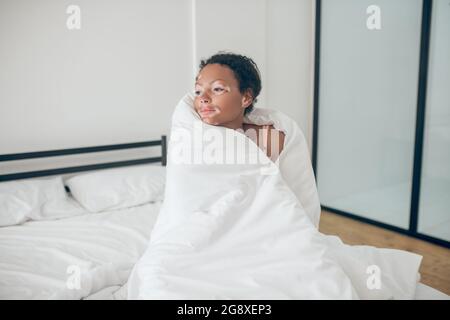 Junge Frau sitzt im Bett mit Decke bedeckt und fühlt sich entspannt Stockfoto