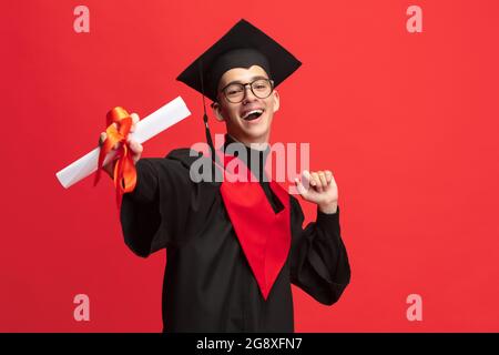 Junge lächelnde, fröhliche Studentin, Absolvent im Mortarboard mit Scroll isoliert über rotem Studiohintergrund. Stockfoto