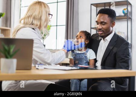 Konzept der Gesundheitsversorgung und Pädiatrie. Rückansicht einer blonden Ärztin, die bei der Generalinspektion den Hals eines Mädchens mit einem Spachtel überprüft. Nettes Mädchen besucht moderne Klinik mit fürsorglichen Vater Stockfoto