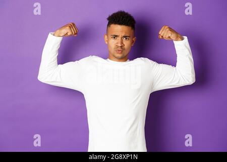 Bild von selbstbewussten afroamerikanischen jungen Kerl, Biegen Bizeps und versuchen zu zeigen, zeigt Muskeln nach dem Training, stehen über lila Hintergrund Stockfoto