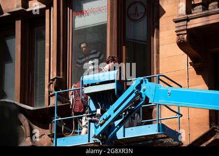 Glasgow, Schottland, Großbritannien. 22. Juli 2021. IM BILD: Ein Fenster bietet dem ortsansässigen Geschäftsmann einen Blick auf das Cherry Picker Team, das die Flaggen aus den Gebäuden entfernt. Die Dekonstruktion der Filmkulisse, die im Stadtzentrum von Glasgow, St. Vincent Street, für die neuen Hollywood-Blockbuster Indiana Jones 5 gedreht wurde. Die Rigger-Crews ziehen ein und nehmen alle amerikanischen Flaggen, Aschen, Ladenschilder und Straßenmöbel herunter und packen sie in Lastwagen. Jedes Set wird dann katalogisiert und gelagert. Glasgow kehrt in eine schottisch aussehende Stadt zurück, nachdem es in den letzten 10 Jahren 1950/60er Jahre in New York war Stockfoto