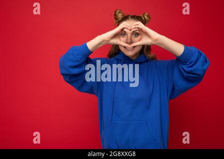 Porträt von jungen positiven schönen blonden Frau mit zwei Hörnern mit aufrichtigen Emotionen tragen lässigen hellblauen Hoodie isoliert auf rotem Hintergrund Stockfoto