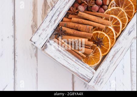 Weiße Holzkiste mit aromatischen Winterwürzen - getrocknete Orangen, Zimtstangen, Anissterne und Nüsse auf Holzhintergrund mit Kopierraum. Stockfoto