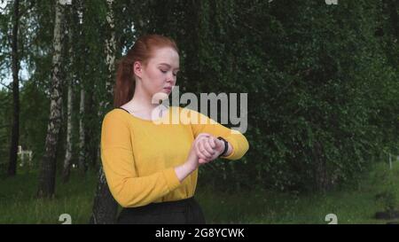 Mädchen mit roten Haaren schaut auf das Armband und hält an, während sie im Park läuft Stockfoto