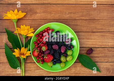 Berry mix - Johannisbeeren, Stachelbeeren, Maulbeeren, Erdbeeren auf einer grünen Untertasse mit gelben Blüten auf einem Holzhintergrund mit Platz für Design. Summe Stockfoto