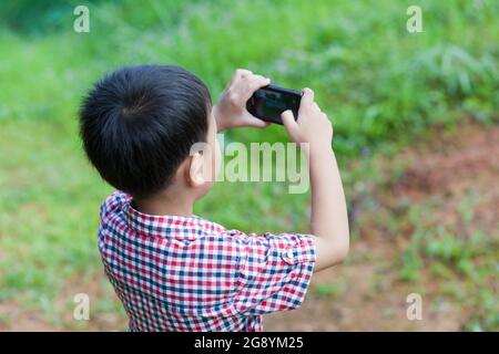 Porträt eines kleinen asiatischen Jungen, der mit guter Absicht Fotos mit der Digitalkamera auf dem Smartphone macht, im Freien inmitten der Lichtung im Urlaub Stockfoto