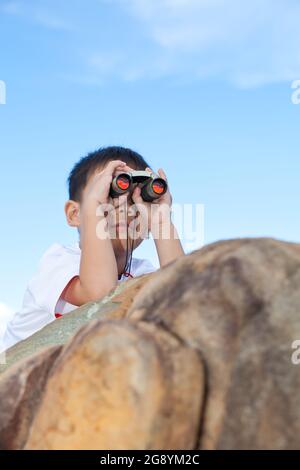 Glücklicher kleiner asiatischer Junge, der mit einem Teleskop auf einem Felsblock im Freien unterwegs ist, und ein kleiner Junge, der mit einem Fernglas den vorderen, hellen Himmelshintergrund betrachtet Stockfoto