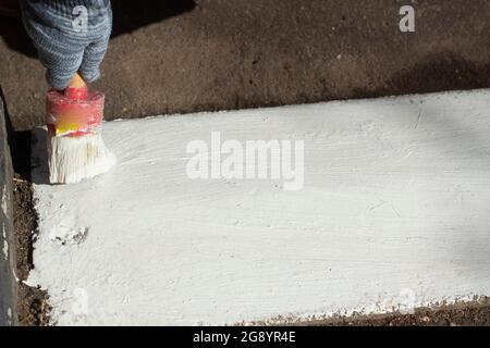 Färbt die Bordsteinkante mit weißer Farbe. Pinsel mit Farbe für die Straße Bordstein. Die Straße lackieren. Der Mann im Handschuh malt. Stockfoto