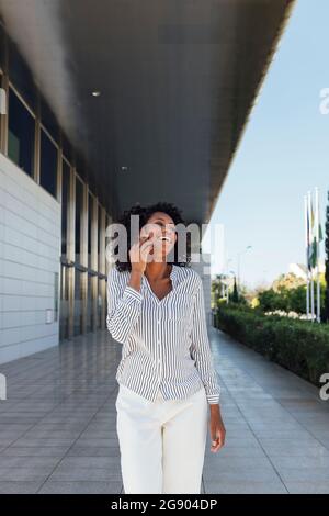 Junge Frau lachte, während sie vor dem Bürogebäude auf dem Mobiltelefon sprach Stockfoto