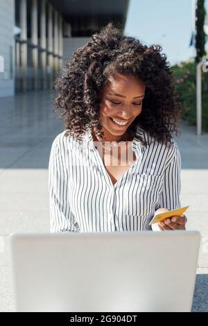 Glückliche junge Geschäftsfrau mit Laptop beim Online-Shopping vor dem Bürogebäude Stockfoto