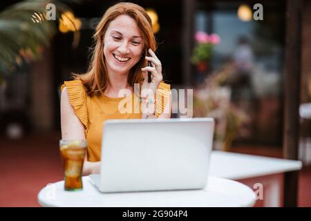 Rotschopf-Frau, die auf einem Smartphone spricht, während sie einen Laptop am Tisch benutzt Stockfoto