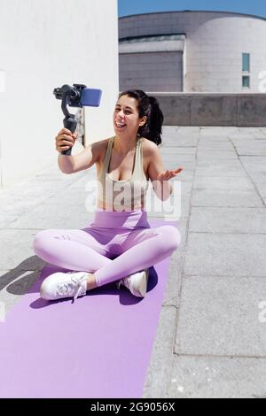 Lächelnde Sportlerin vloggt durch das Smartphone, während sie an sonnigen Tagen auf der Trainingsmatte sitzt Stockfoto