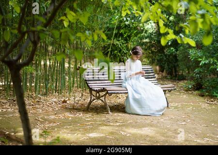 Mädchen in weißem Abendkleid sitzt auf der Bank im Park Stockfoto