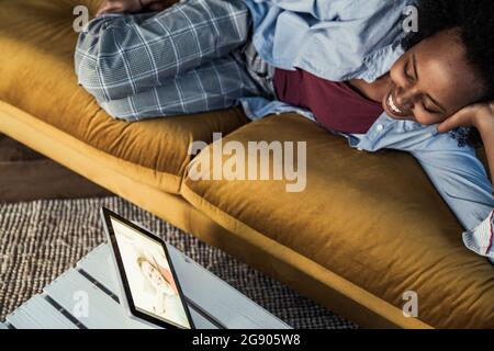 Frau, die mit einer Freundin über ein digitales Tablet spricht, während sie auf dem Sofa liegt Stockfoto