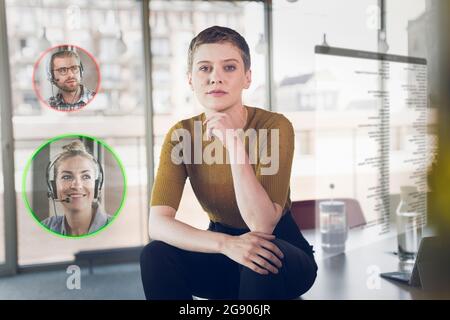 Geschäftsfrau mit Hand am Kinn auf dem Schreibtisch mit Ikonen von männlichen und weiblichen Kollegen in der Ecke Stockfoto