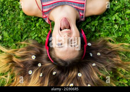 Schelmische junge Frau, die aus der Zunge ragt, mit Blumen im Haar, die auf dem Gras liegen Stockfoto