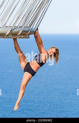 Glückliche, reife Frau, die an einem Geländer hängt und sich das Bein streckt, während sie am Aussichtspunkt Akrobatik übt Stockfoto