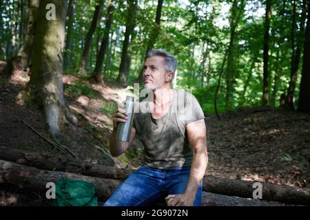 Mann, der aus einer Wasserflasche trinkt, sitzt auf einem Baumstamm im Wald Stockfoto
