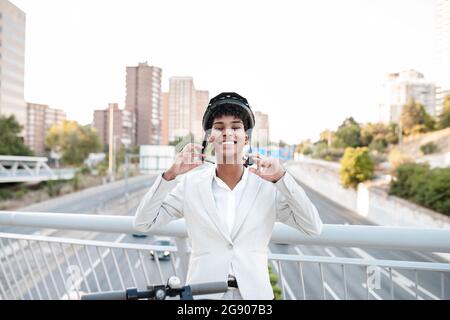 Glückliche professionelle Frau trägt Helm, während sie auf der Brücke in der Stadt steht Stockfoto