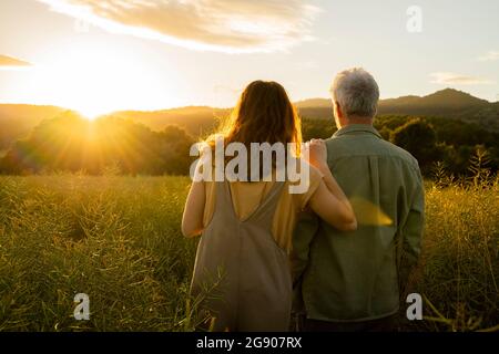 Tochter und Vater blicken auf den Sonnenuntergang, während sie auf dem Feld stehen Stockfoto