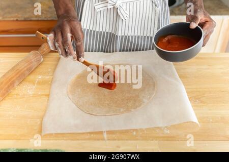 Mann, der Tomatensauce in der Küche zu Hause auf Pizzateig legt Stockfoto