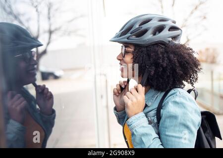 Lächelnde junge Frau, die beim Tragen des Fahrradhelms auf das Glasfenster blickt Stockfoto