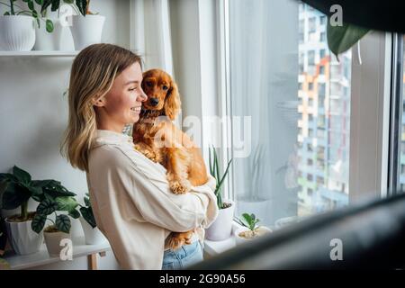 Lächelnde junge Frau, die den Hund Cocker Spaniel trägt, während sie zu Hause durch das Fenster schaut Stockfoto