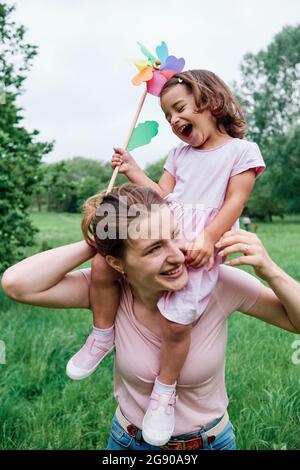 Lachende Tochter hält das Windrad, während sie auf der Schulter der Mutter im Park sitzt Stockfoto