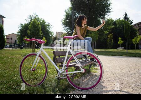 Junge Frau mit Fahrrad, die Selfie über das Smartphone nimmt, während sie auf der Bank sitzt Stockfoto