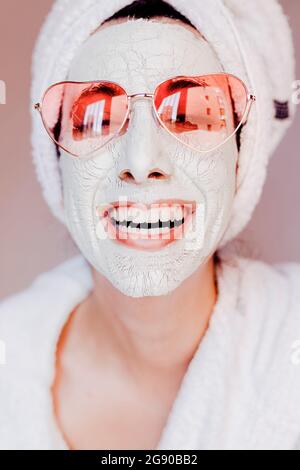 Fröhliche junge Frau mit Beauty-Gesichtsmaske trägt herzförmige Sonnenbrille Stockfoto