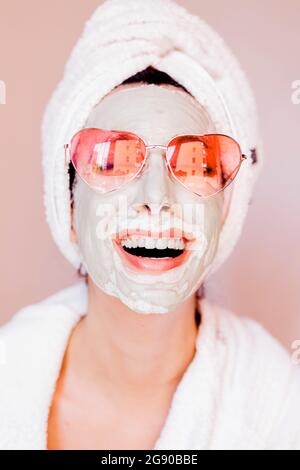Glückliche junge Frau mit Beauty-Gesichtsmaske trägt herzförmige Sonnenbrille Stockfoto