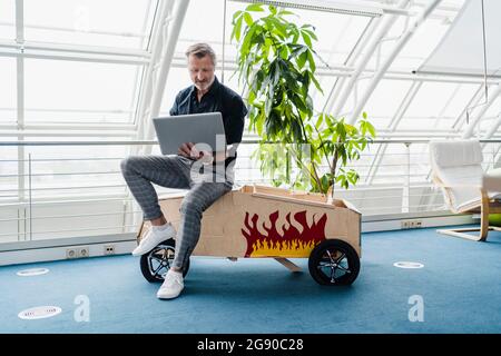 Geschäftsmann mit Laptop, während er im kreativen Büro auf einem Spielzeugauto sitzt Stockfoto