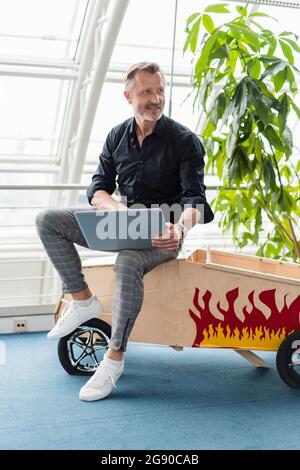 Lächelnder Geschäftsmann, der im Büro mit einem Laptop auf einem Autospielzeug sitzt Stockfoto