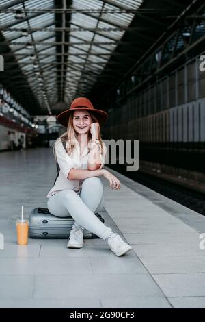 Eine glückliche Frau mit Hut sitzt auf dem Gepäck am Bahnhofsplatz Stockfoto