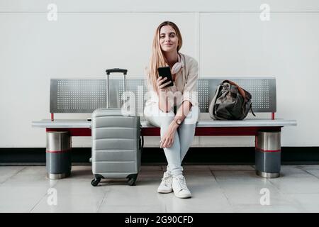 Schöne Frau hält Smartphone, während sie mit Gepäck auf Rädern am Bahnhof sitzt Stockfoto