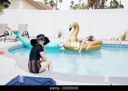 Lächelnde Frau am Pool, während sich der Mann am sonnigen Tag auf dem aufblasbaren Schwan entspannt Stockfoto