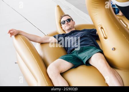 Ein hübscher Mann mit Sonnenbrille, der auf dem schwimmenden Wagen auf der Terrasse liegt Stockfoto